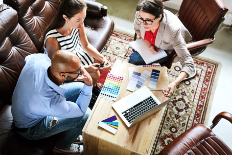 Three people are shown collaborating around a small table, reviewing color swatches and design plans.