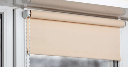 A tan roller shade is mounted on a white window frame.