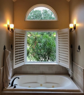 A bathroom features a white tub, open white shutters, and two wall sconces.