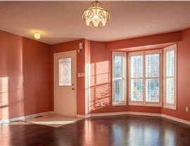An interior view showcasing a window replacement, featuring a bay window with white shutters in a room with salmon-colored walls and a dark wood floor.