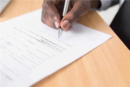 Close-up of a hand signing a contract on a wooden desk, the document appears to be a purchase agreement.