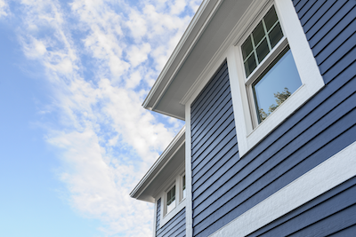A low-angle shot of blue siding with white trim and windows against a partly cloudy sky.