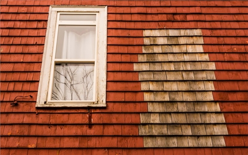 A close-up shows a section of a house with red wooden shingles and a white-framed window on the left, along with a lighter-colored area of shingles on the right indicating siding replacement.