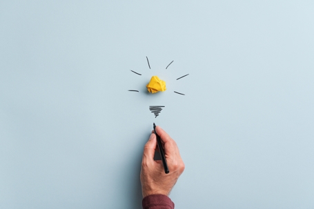 A hand draws the base of a lightbulb, with a crumpled yellow paper acting as the top, on a pale blue background.