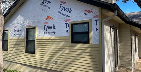 Residential home exterior showing construction in progress, featuring Tyvek HomeWrap, yellow siding, and black window trim.