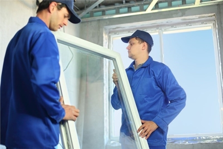 Two workers in blue uniforms carry a window frame during installation.