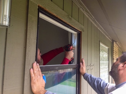 Two people install a window. One holds the frame while the other uses a drill to secure it.