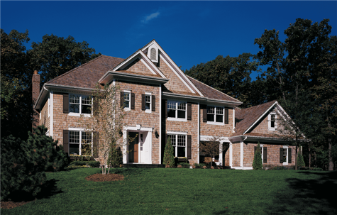 A two story home with brown shingles and white windows sits on a lush green lawn, surrounded by tall trees.