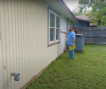 A man stands outside a home and points to a window while holding a clipboard.