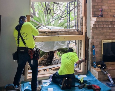 Two workers in yellow shirts, "You & Service with Love & Excellence", are installing a new window, with tools at their feet.