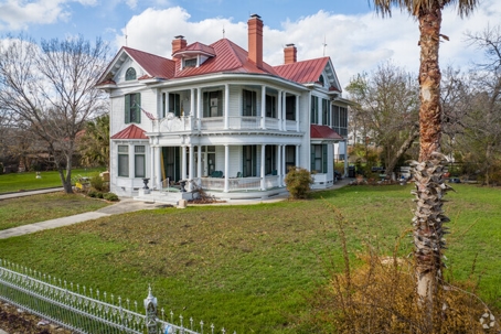 two-story white Victorian home with a red roof, a manicured lawn, and a decorative fence.