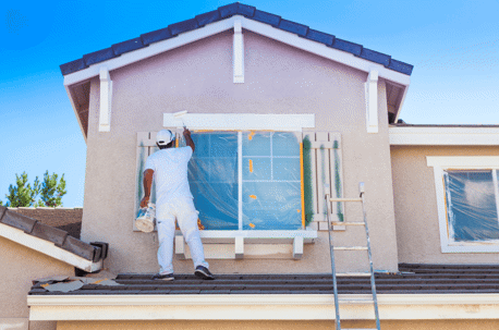 An exterior house painter applies white paint to a window frame of a home.