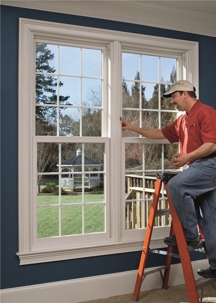 A man on a ladder adjusts a new double hung window, with a backyard and gazebo visible through the glass.