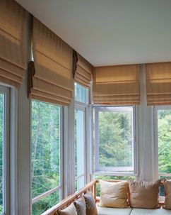 A sunroom with tan Roman shades and white framed windows looks out onto a green forest.