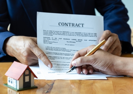 Close-up of two hands reviewing a contract on a wooden table, next to a small model house.