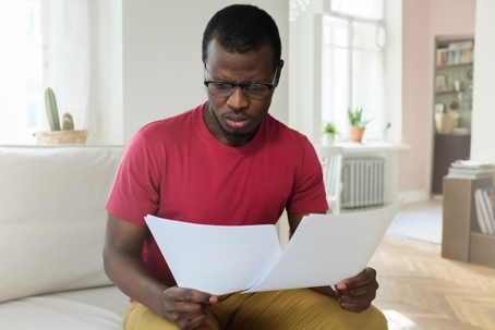 A man in a red shirt appears concerned as he reviews documents in his home.