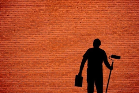 A silhouette of a person using a paint roller is cast against a red brick wall, demonstrating an action.