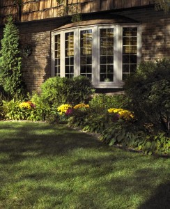 A sunlit lawn leads to a brick house with a large bay window.