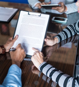 Two people review a contract document held by one, with another using a pen to note something.