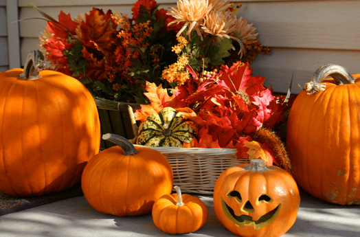 Orange pumpkins and jack-o-lanterns with fall foliage sitting on a porch front. 