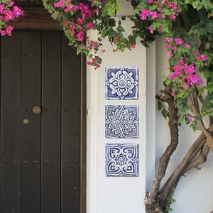 Three small square blue and white ceramic exterior wall decor hanging from a white stucco home with a brown door next to it and a small tree with pink blossoms to the right.