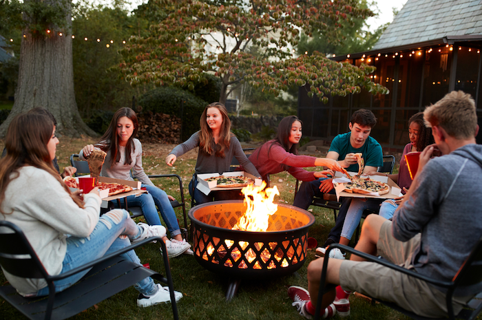 A group of millennial-aged men and women sitting around a fire pit sharing pizza during the evening time.