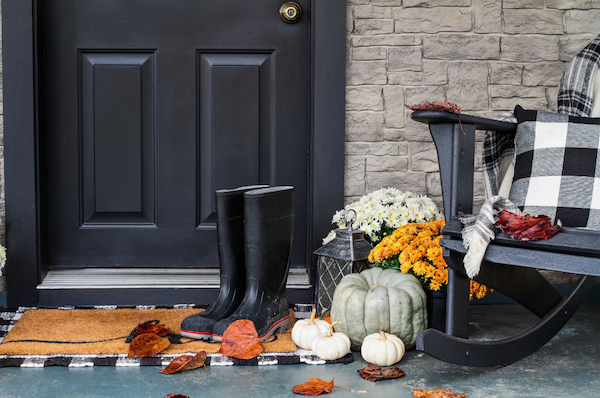 The front entry way of a home with a black rocking chair, blanket, and pillow with fall pumpkins, mums, and black rain boots at the foot of the front door.