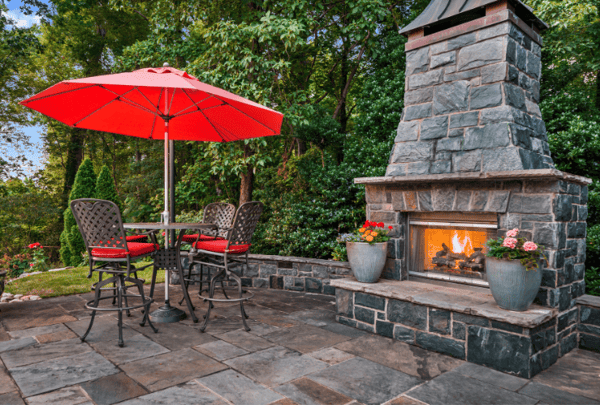 A red umbrella over an outdoor dining set on a patio with a brick fireplace and trees behind them. 