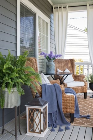 A front porch of a home with two whicker chairs with white pillows and a blue blanket on one chair. There is a fern plant to the left of a chair and a floor lantern with a large brown patterned rug underneath the chairs.