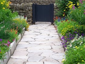 A sidewalk made of landscaping stones surrounded by bright colorful flowers and greenery leading up to a black gate.