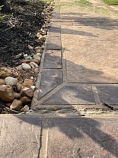 A close up of a sidewalk with stamped concrete detailing that looks like stone. There are small rocks and plants surrounding the sidewalk. 
