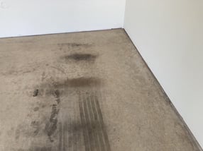 The corner of an empty garage floor with a smooth surface and white walls. There are some stains on the concrete coating.