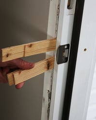 A close up shot of a door frame where the closing mechanism is. There are skinny wooden pieces wedged between the door frame and wall, or jamb, and a hand holding one side of the wedges.