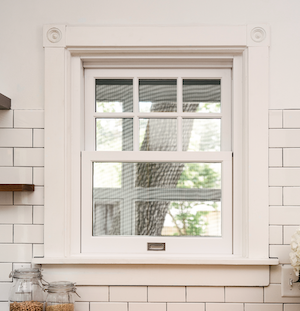 A small single-hung window in a kitchen with white backsplash and a white frame with a grid pattern on the bottom sash.
