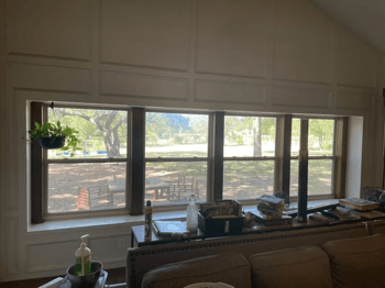 A room features a large window with brown trim. The window looks out onto a backyard with trees, a f…