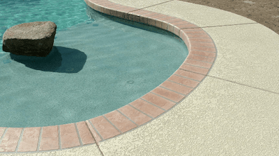 The corner of a rounded swimming pool with red tile around the pool and a beige colored concrete wit…