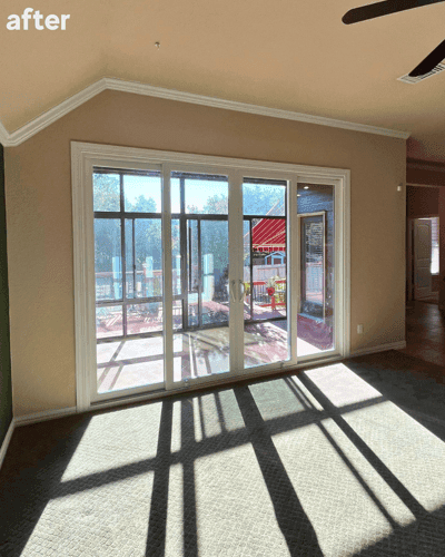 A white sliding patio door in place of the three windows in the same living room with light streamin…