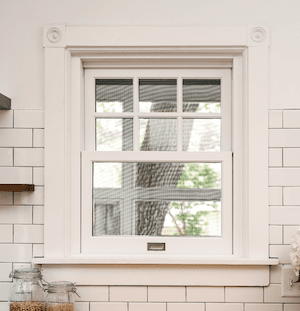 A small single-hung window in a kitchen with white backsplash and a white frame with a grid pattern…