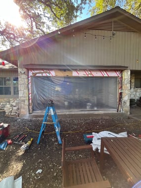 The exterior of a home with the four windows removed and a large opening covered by a plastic tarp where the new door will be placed. 