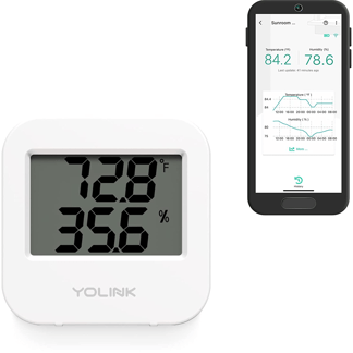 A white digital thermometer that reads temperature and humidity levels with a smartphone next to it. 