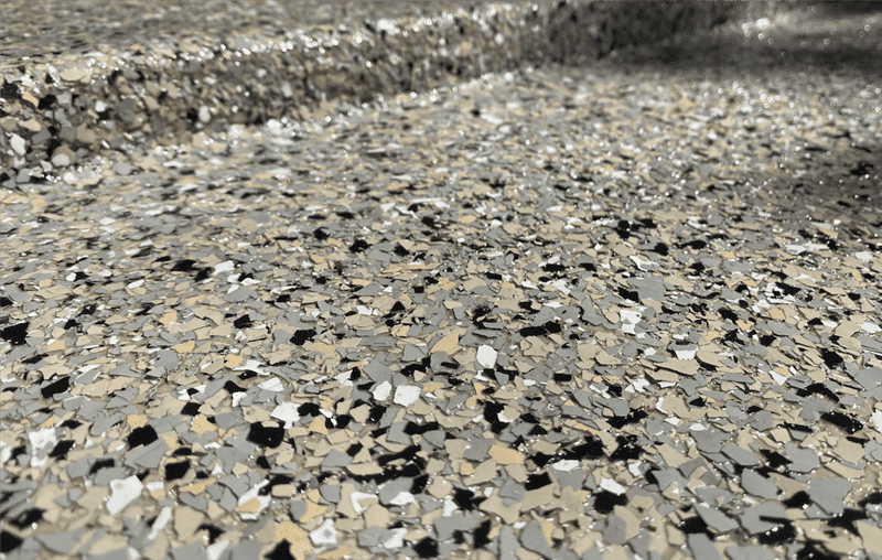 A close-up of a garage floor with a Penntek concrete coating that is made up of beige, white, grey, and black small vinyl chips with a glossy topcoat.