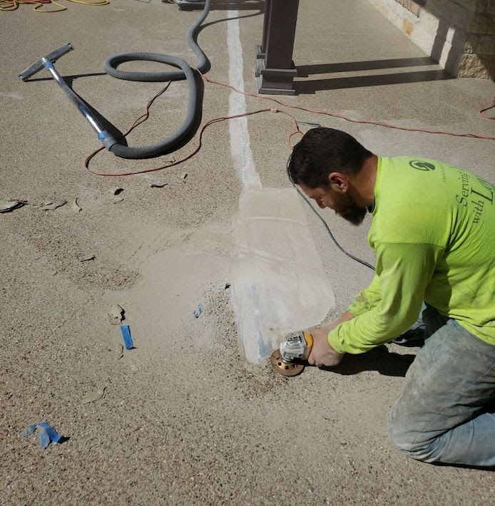 A man in a bright yellow long sleeve shirt on his knees on a back patio with a small hand grinder to the concrete patio, smoothing out the surface. There is a vacuum hose in the background.