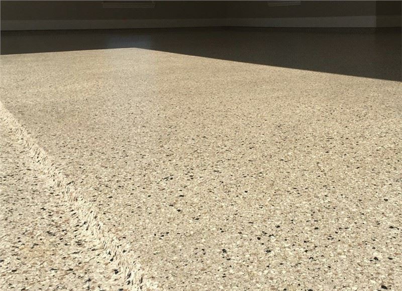 A close up of a garage floor apron and lip with a concrete coating that looks like granite with tan,…