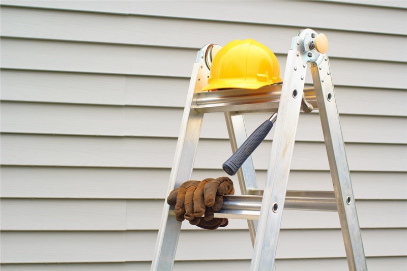 A silver ladder with work gloves, a hammer, and yellow hardhat on it sitting in front of the side of a home with lap siding.