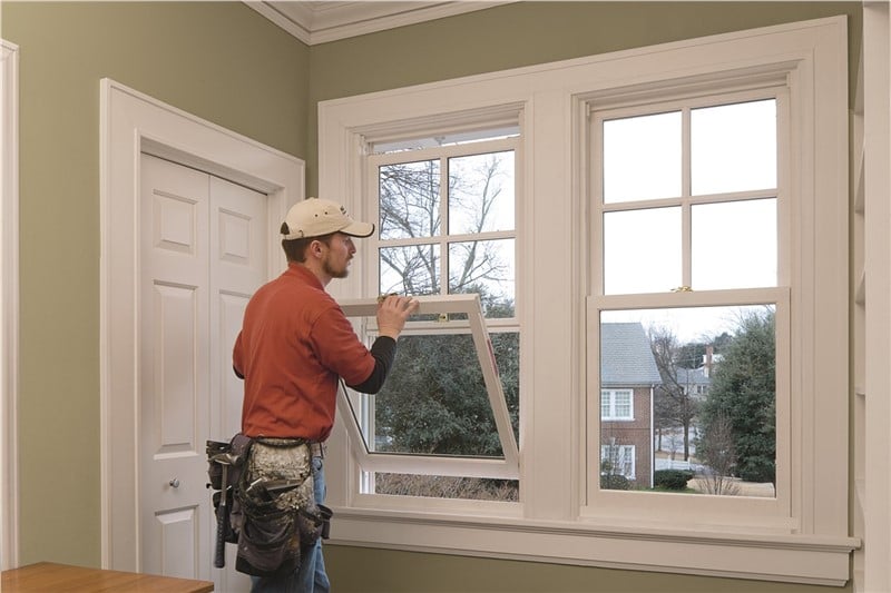 A man in an orange shirt and tan baseball cap opening the bottom sash of a single hung window. of the inside of a home.