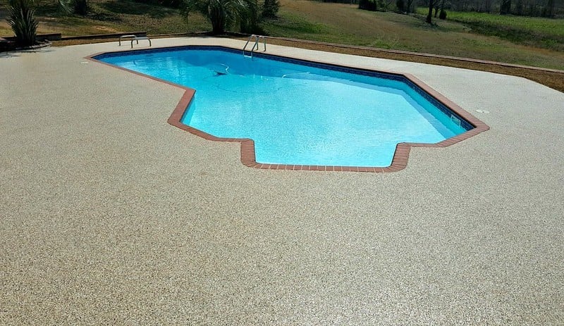 A clear blue pool that is a geometric shape with a pool deck coating surrounding that is a tan color with multicolor chips. There is green grass in the background.
