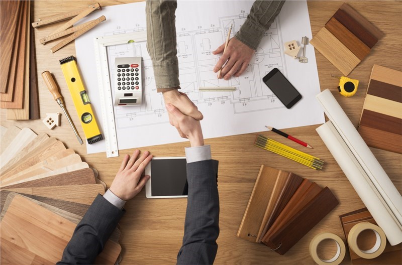 A stock image of two businessmen shaking hands across a table. There are design plans on the table and wood samples surrounding them.