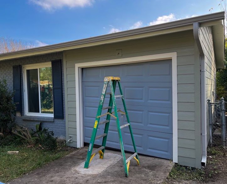 The outside of a home with a small blue garage door and a toned-down green siding color. There is a bright green ladder in front of the garage door and the blue sky in the background.