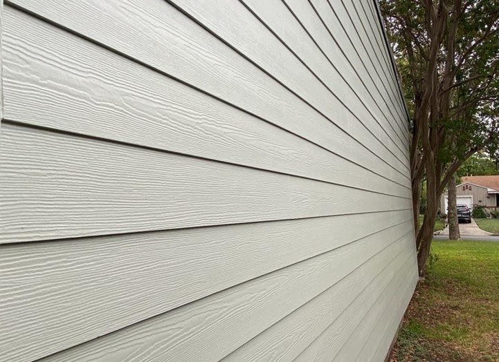 The side of a home with beige horizontal lap siding  with grass and a small tree in the background.