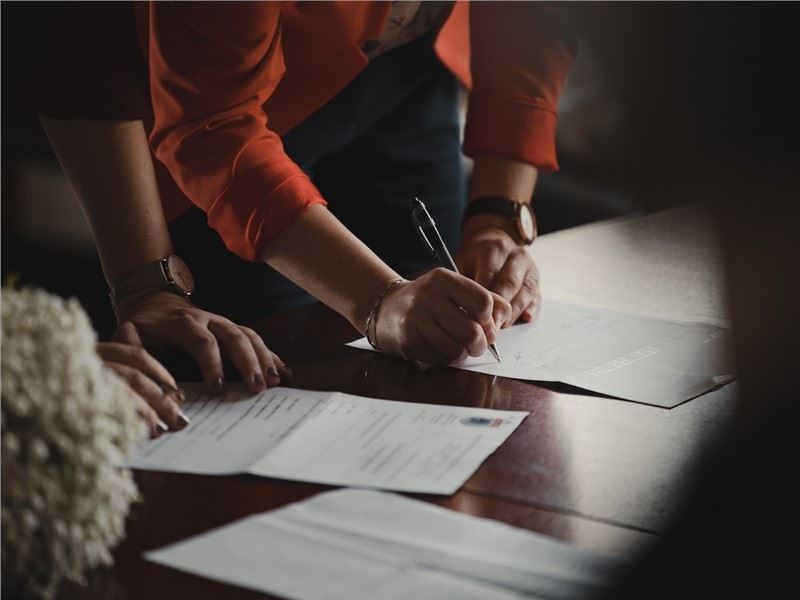 Viewed from over someone's shoulder, the arms of two people leaning against a table looking at paper…